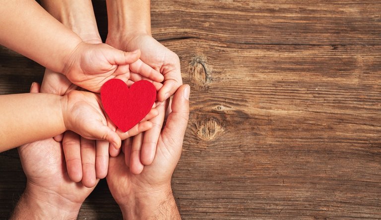 Zwei Erwachsenen Hände und eine Kinder Hand halten ein rotes Herz vor einem Holzhintergrund
